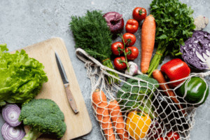 Fabric bag of vegetables on a table next to a cutting board.