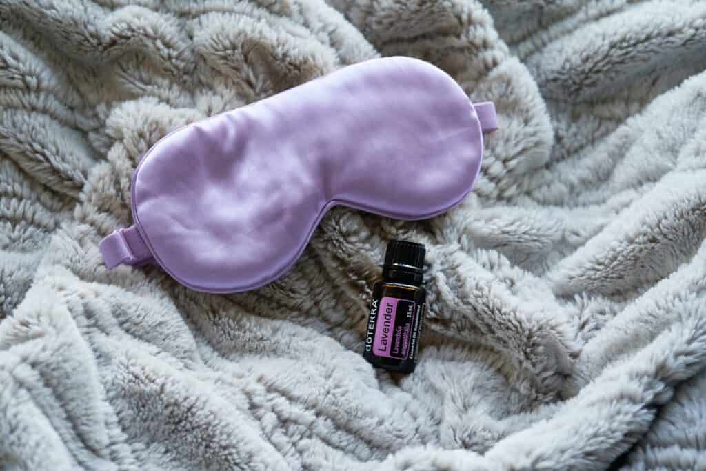 Lavender essential oil bottle next to an eye mask on a soft gray blanket.