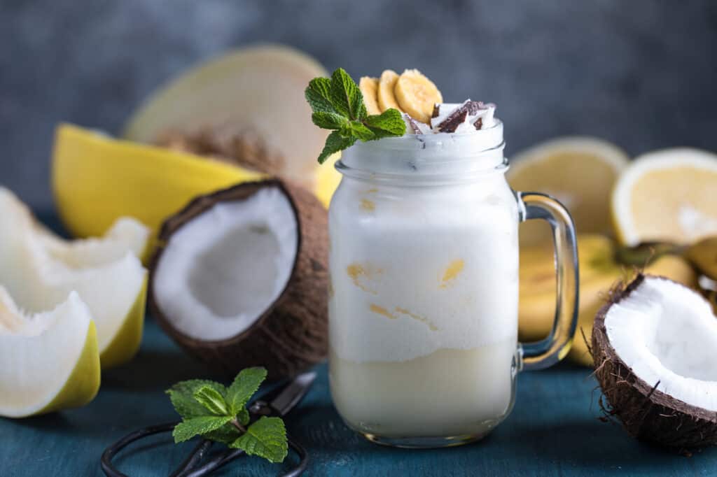 Fresh summer pina colada cocktail with exotic fruits: banana, melon, coconut milk, pineapple juice, white grapefruit, lemon and mint in the jar on the blue wooden background